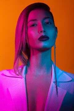 In the club concept. Fashion portrait of rich glamourous woman wearing luxurious earrings, posing over orange background. Pink, blue neon lights. Close up. Synthpop style. Studio shot