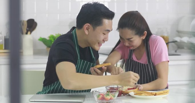 Lovely Asian couple spread strawberry jam on bread together, man bait woman, concept for lovely Asian couple in kitchen.