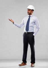 architecture, construction business and building - smiling indian architect or businessman in helmet showing something imaginary over grey background