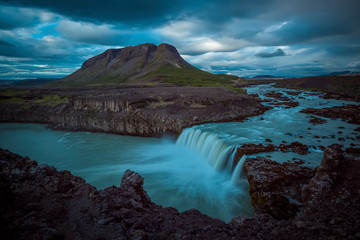 The Bjófafoss Waterfall with golden clouds in the sky. The flowing water is captured by a long...