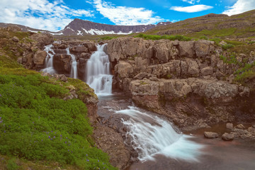 Fototapeta na wymiar The Noname Waterfall with golden clouds in the sky. The flowing water is captured by a long exposure. Amazing blue color of water from the glacier. Natural and colorful environment...