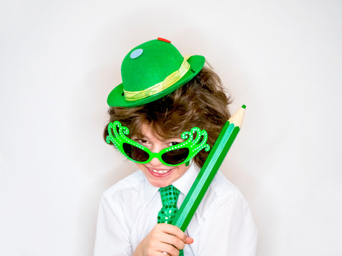 Funny St Patricks day stunning little boy wearing a green hat, carnival glasses and a tie. Curly child posing with a big green pencil in hand on a light background