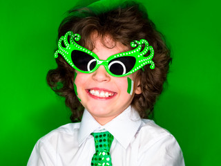 Child Celebrating St. Patrick's Day Showing his Make-up. A small, curly smiling boy in green carnival accessories looking at the camera. green background