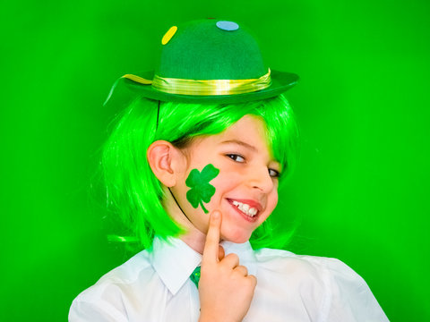 Child Celebrating St. Patrick's Day Showing his Make-up. A small, boy in green carnival wig points his finger at a drawing in the form shamrock on his cheek.green background