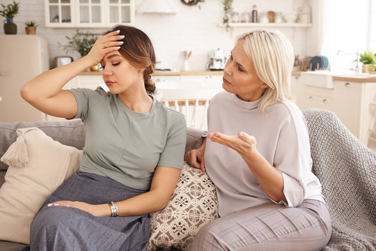 Picture of worried mature blonde woman trying to have conversation with her annoyed upset daughter, asking what's wrong. Beautiful young female ignoring her mother while having disagreement