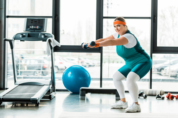 cheerful plus size woman exercising with dumbbells near treadmill