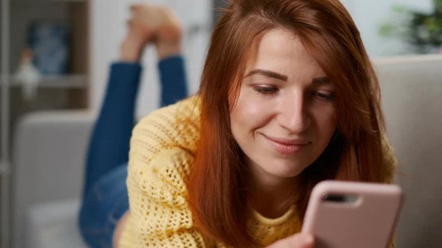 Portrait of young single woman using dating app on her smartphone. Girl in the yellow sweater holding a smartphone in hands and typing on it chatting in the living room. Lovely home atmosphere.