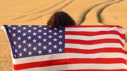 4K video clip of mixed race African American girl teenager female young woman holding an American USA Stars and Stripes flag in a wheat field at sunset or sunrise