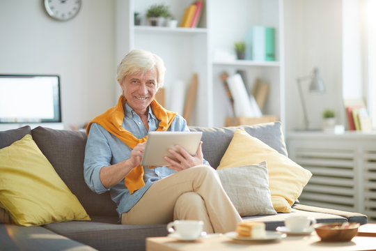 Portrait of contemporary senior man smiling happily while using digital tablet sitting on sofa at home, copy space