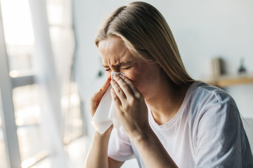Sick yong blonde woman sneeze into white napkin. She sit in room at window. Model suffer. She feels bad