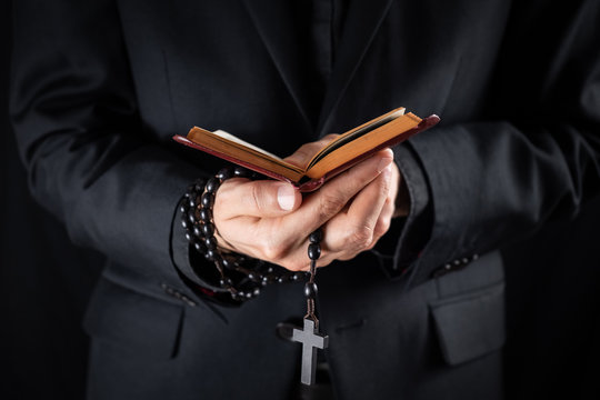 Hands of a christian priest dressed in black holding a crucifix and reading New Testament book. Religious person studies Bible and holds prayer beads, low-key image