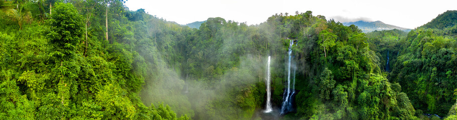 Aerial over Sekumpul waterfall surrounded by dense rainforest and mountains shrouded in mist at sunrise, Bali, Indonesia panoramic