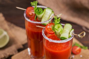 Two glasses of tomato juice decorated with fresh tomatoes, cucumber and leaves on a wooden background