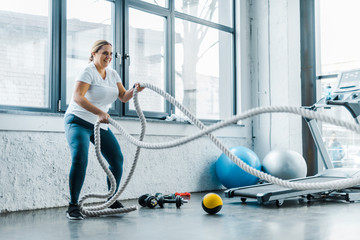 cheerful overweight woman training with battle ropes in gym