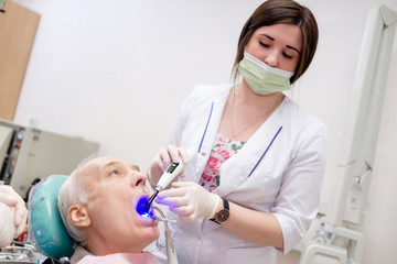Dentist woman working with dental polymerization lamp in oral cavity of senior man patient. Dental care for elder people. Dentist holding dental device. Dental care for elder people.