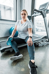 tired plus size woman resting on treadmill and holding bottle with water