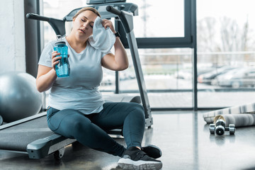 tired plus size woman wiping sweat with towel while sitting on treadmill and holding bottle with...