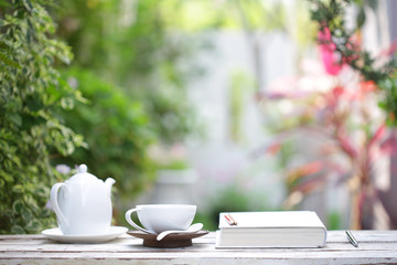 White coffee cup and kettle with notebooks on wooden table with green plants