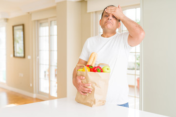 Middle age man holding groceries shopping bag at home stressed with hand on head, shocked with shame and surprise face, angry and frustrated. Fear and upset for mistake.