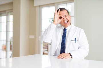 Fototapeta premium Middle age doctor man wearing medical coat at the clinic doing ok gesture with hand smiling, eye looking through fingers with happy face.