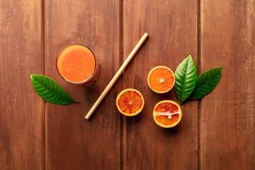 An overhead photo of a glass of orange juice with blood oranges, green leaves, and a bamboo straw