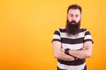 Handsome bearded man with stripped t-shirt holding arms crossed and looking at the camera over yellow background