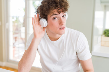 Young man listening to music wearing headphones at homes smiling with hand over ear listening an hearing to rumor or gossip. Deafness concept.