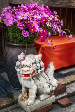 Small lion-dog stone statue in front of a large vase with flowers. Lion-dogs or Komainu are the guardians of the gates to shrines and houses.