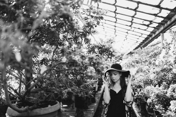 Young beautiful caucasian woman in glass greenhouse among colorful azalea flowers. Art portrait of a girl wearing a hat and romantic boho clothes. Gardening, plant growing, art therapy concept.