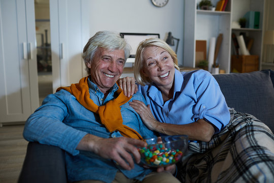Portrait of laughing mature couple watching tv in dark sitting on comfortable sofa, copy space