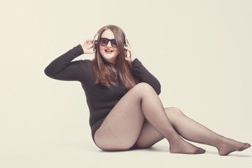 Naklejka premium Model plus size with sweet donut, happy girl posing with headphones and listen music, in black bodysuit. XXL female sitting on light background