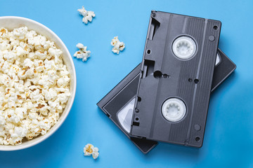 Two black VHS tapes, bowl of popcorn and scattered popcorns on a blue background. Movie/ film industry. Retro/ vintage technology. Entertainment.