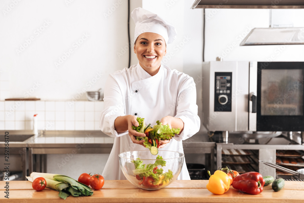 Wall mural photo of adult woman chief wearing white uniform making salad with fresh vegetables, in kitchen at t