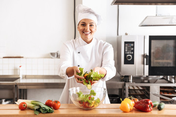 Photo of adult woman chief wearing white uniform making salad with fresh vegetables, in kitchen at the restaurant