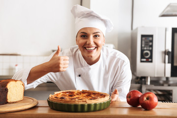 Image of joyful woman chef wearing white uniform, posing in kitchen at the cafe with baked goods