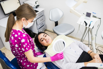 Smiling girl with a mirror in the right hand on the patient chair in the dental cabinet.