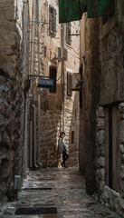 A girl stands in a narrow street in an old European city with a cobbled road and stone walls.
