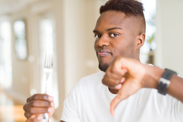 African american man holding silver fork cutlery with angry face, negative sign showing dislike...