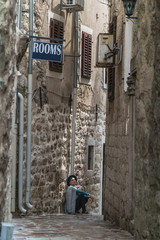 A girl stands in a narrow street in an old European city with a cobbled road and stone walls.