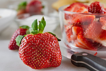 Close up view of a strawberry on a white cutting board with part of a knife.  Additional strawberries already cut in bowl to the side.  Blurry background with bowls of sugar and pastries.