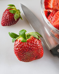 Close up of strawberries and a knife on white cutting board.  Partial bowl of already sliced berries in upper right corner.