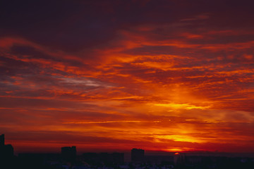 Cityscape with vivid fiery dawn. Amazing warm dramatic cloudy sky above dark silhouettes of city buildings. Orange sunlight. Atmospheric background of sunrise in overcast weather. Copy space.