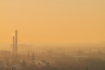 Smog among silhouettes of buildings on sunrise. Smokestack in dawn sky. Environmental pollution on sunset. Harmful fumes from stack above city. Mist urban background with warm orange yellow sky.