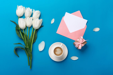 White tulips with petals, cup of coffee, gift box, a love note and envelope on a blue background