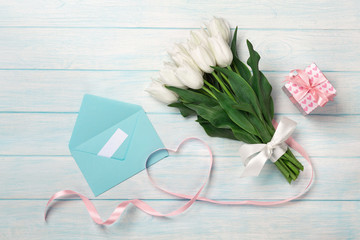 A bouquet of white tulips and a pink ribbon in the form of a heart with a gift box, love note and color envelope on blue wooden boards