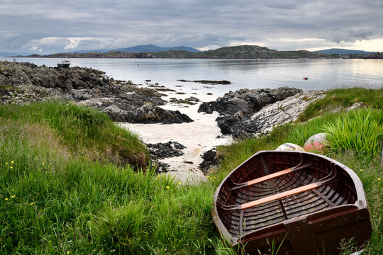 Rocky shore of Isle of Iona with beached boat with Fionnphort Isle of Mull and mountains of Ben More past Sound of Iona Inner Hebrides Scotland UK