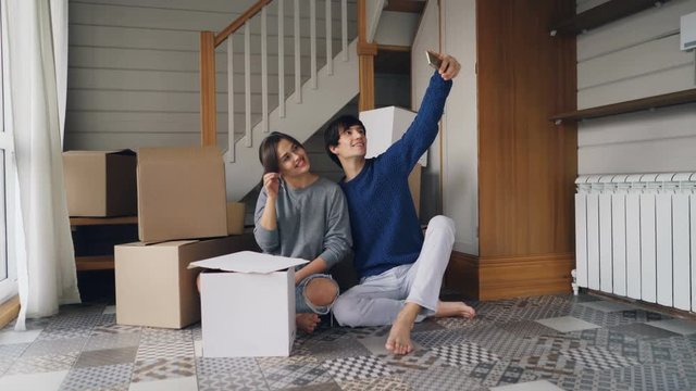 Happy family husband and wife are taking selfie with house keys sitting on floor of new apartment near carton boxes with things. Relocation and social media concept.