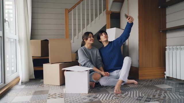 Adorable young people girl and guy are taking selfie expressing love and tenderness kissing and hugging sitting on floor of new house and using smartphone.