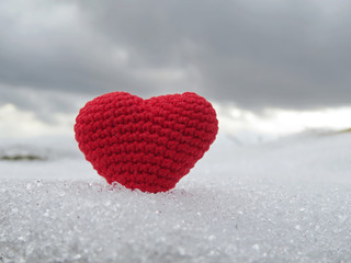 Fototapeta premium Red knitted heart in the snow on cloudy sky background, symbol of pure love. March 8, Valentine day, concept of romantic holiday, health care or blood donation