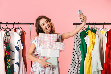 Photo of elegant woman with purchase standing in store near clothes rack and taking selfie on cell phone